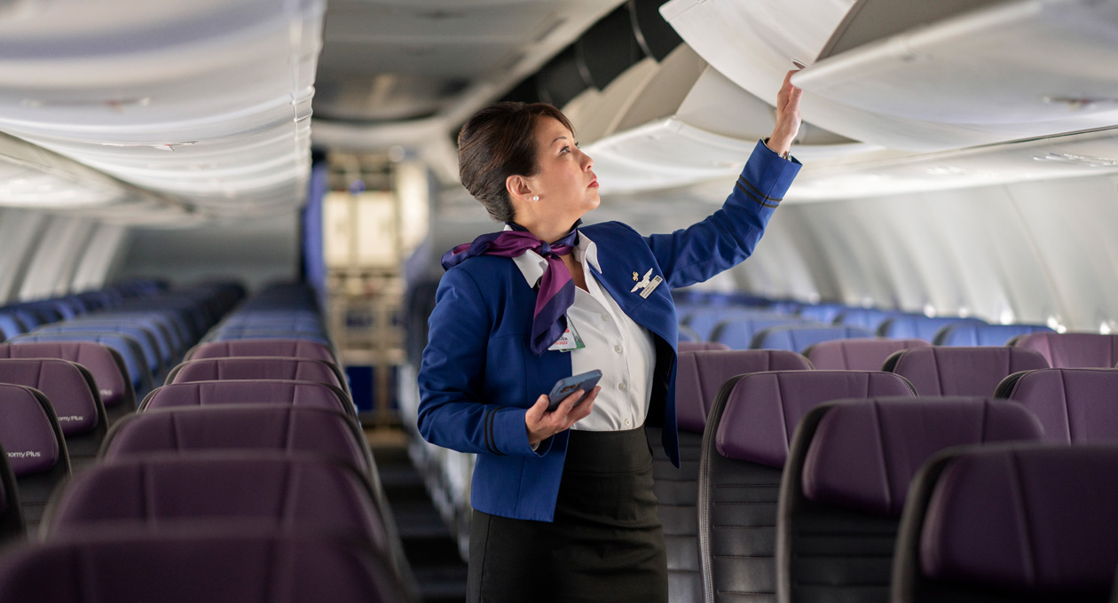 在空無一人的機艙內，一名空中服務員手持 iPhone 並同時打開上方行李架