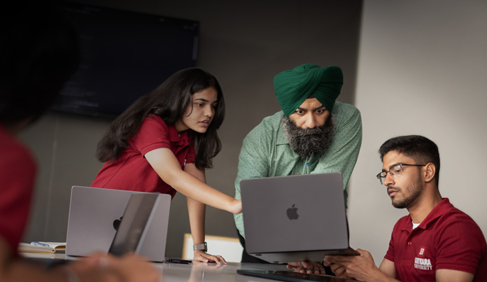 A professor at a classroom table leans over to look at his student’s MacBook screen, while another student points out something on the screen.