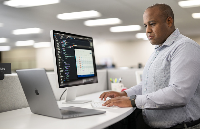Um desenvolvedor está à mesa usando um Mac e um MacBook com código de programação e informações do app mostrados na tela do Mac