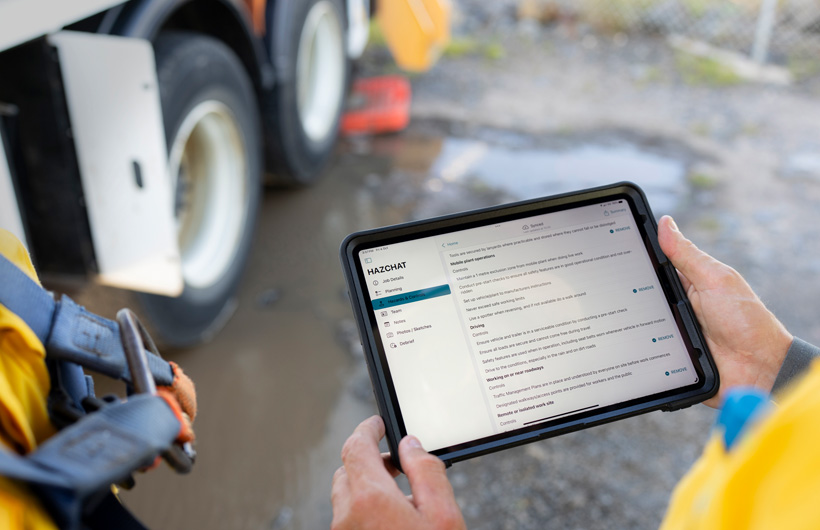 Deux mains tiennent un iPad avec l’app HazChat ouverte affichant « Job Details », « Planning », « Hazards » et « Controls », tandis qu’une personne est sur un chantier près d’une flaque de boue