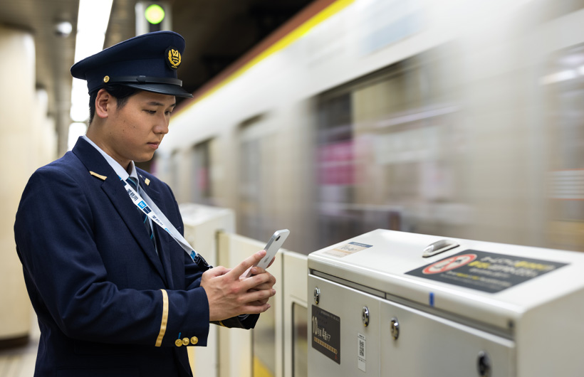 Un agent de station se tient sur le quai et utilise son iPhone pour scanner un code QR tandis qu’un métro passe.
