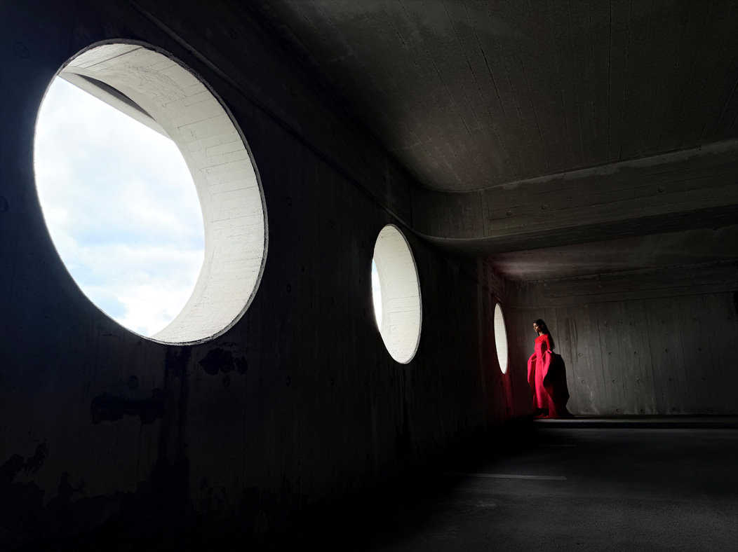 Une femme vêtue d’une longue robe rouge se fait photographier dans une pièce vide. L’app Appareil photo change de distance focale pour illustrer les capacités du système caméra pro, mettant en valeur : 24 mm